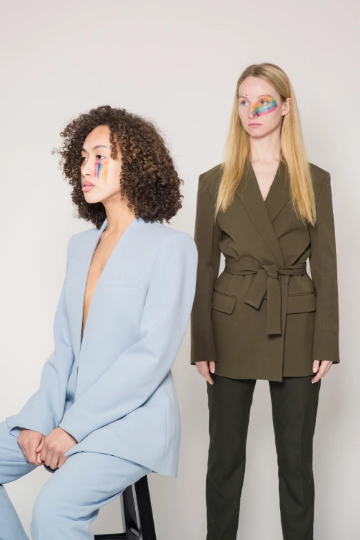 two women standing next to each other in front of a white wall, inspired by Vanessa Beecroft, trending on unsplash, color field, wearing a light blue suit, green and brown clothes, pale and coloured kimono, one blonde and one brunette