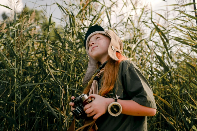 a woman standing in tall grass holding a camera, a picture, trending on pexels, girl wearing uniform, kid, wearing a fisher 🧥, wearing adventure gear