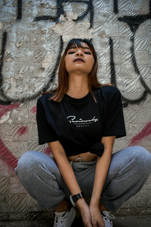 a woman sitting on the ground in front of a graffiti wall, a cartoon, by Robbie Trevino, trending on unsplash, renaissance, wearing a black!! t - shirt, official product photo, phantasy, rembranlt