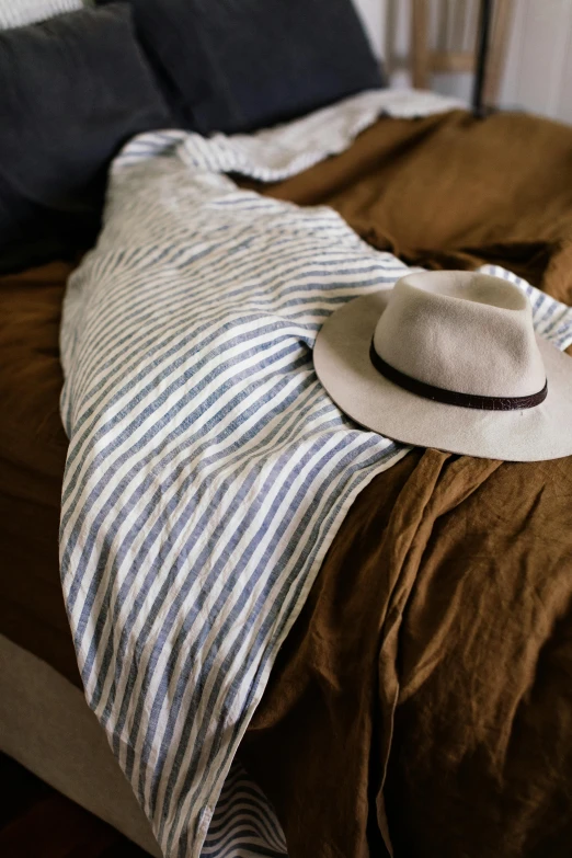 a hat sitting on top of a bed next to pillows, a portrait, by Nina Hamnett, unsplash, striped shirt, burnt umber and blue, cloth wraps, linen