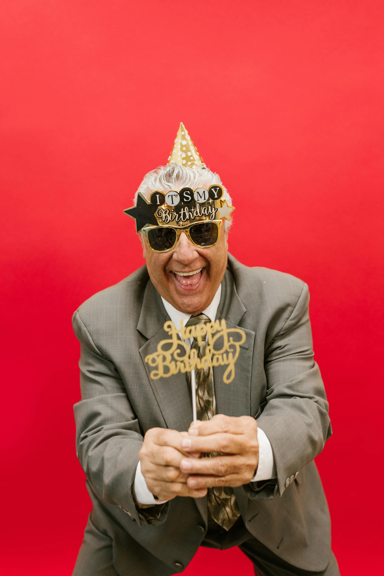 a man wearing a party hat and holding a cake, an album cover, by Bernie D’Andrea, pexels, silver silver glasses, kevin o'leary, old man, gold suit