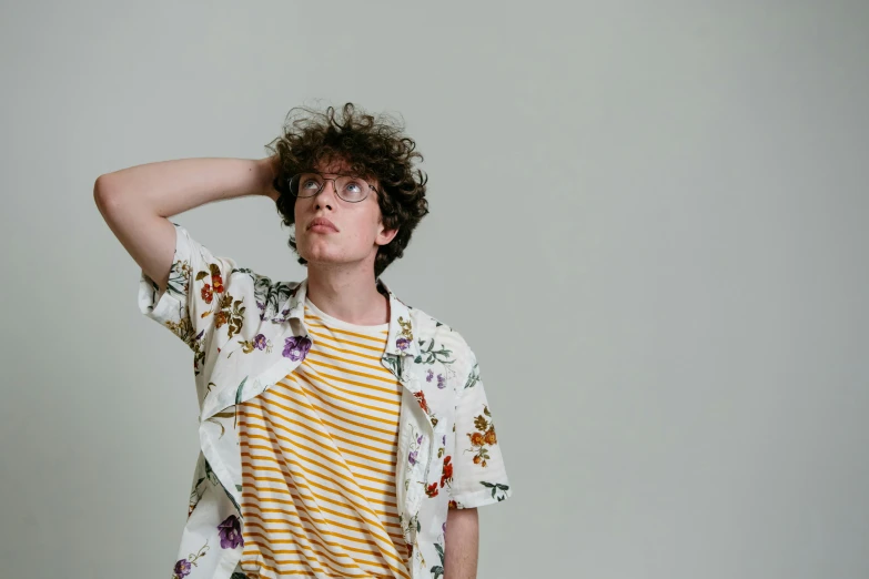 a young man standing in front of a white wall, an album cover, by Carey Morris, trending on pexels, wearing yellow floral blouse, curly haired, jewish young man with glasses, thinking pose