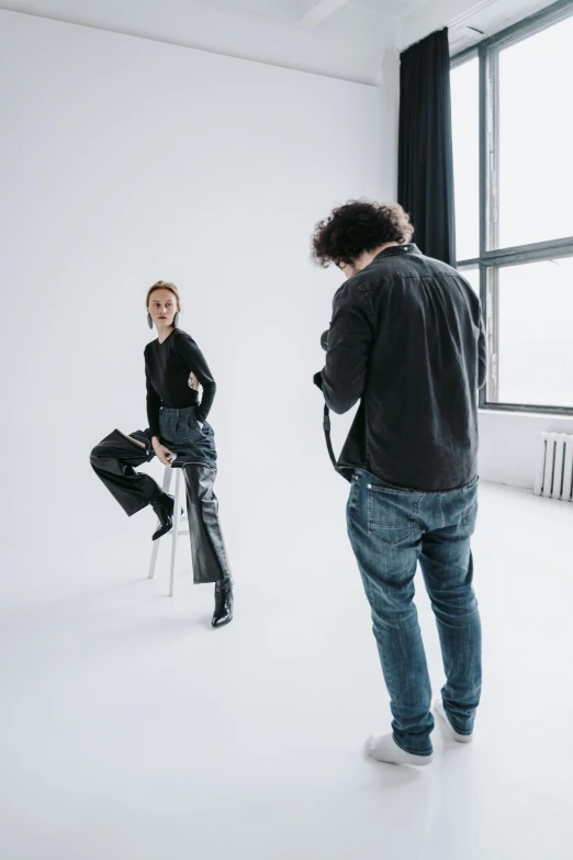 a man taking a picture of a woman in a white room, inspired by Peter Lindbergh, trending on unsplash, jeans and knee high black boots, sitting on designer chair, studio backdrop, black leather garment