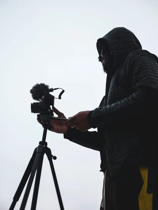 a man standing next to a camera on a tripod, unsplash, video art, on a cloudy day, ( ( theatrical ) ), holding a microphone, photography )