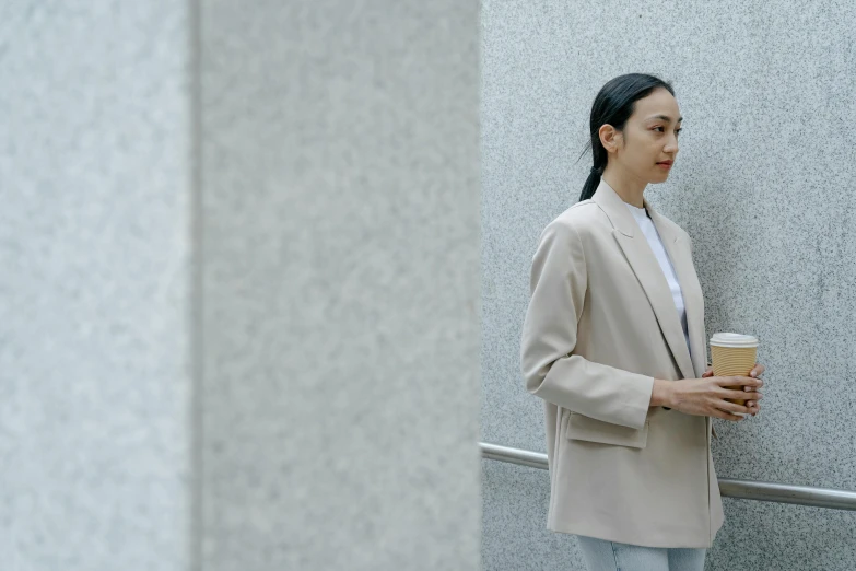 a woman standing next to a wall holding a cup of coffee, trending on pexels, neo-figurative, tan suit, ethnicity : japanese, grey jacket, cinematic outfit photo