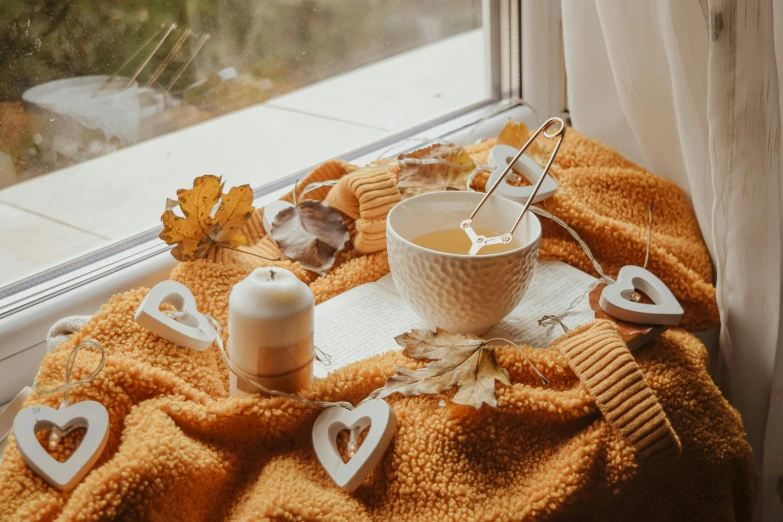 a cup of coffee sitting on top of a table next to a window, a still life, inspired by Elsa Bleda, trending on pexels, 🍂 cute, floating candles, orange and white color scheme, covered with blanket