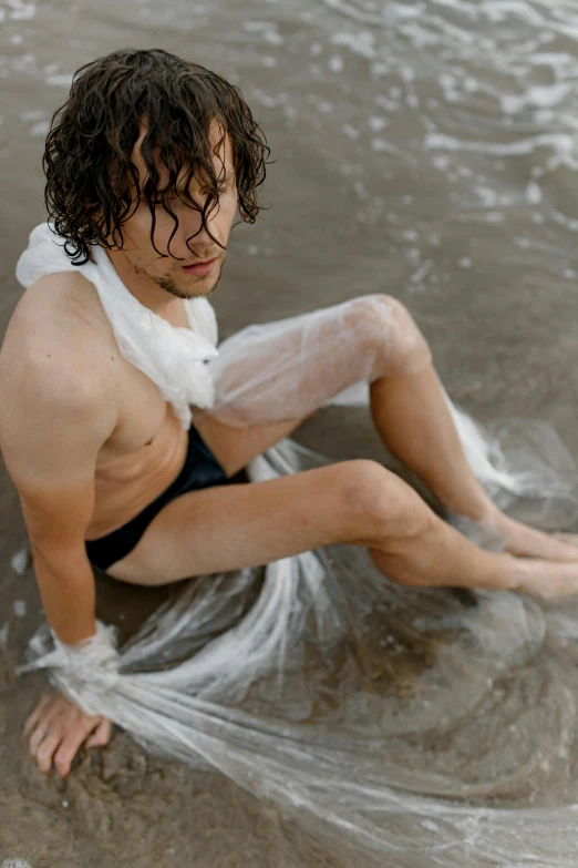 a man sitting on top of a beach next to the ocean, renaissance, wet skin and windblown hair, wearing translucent sheet, non binary model, curls