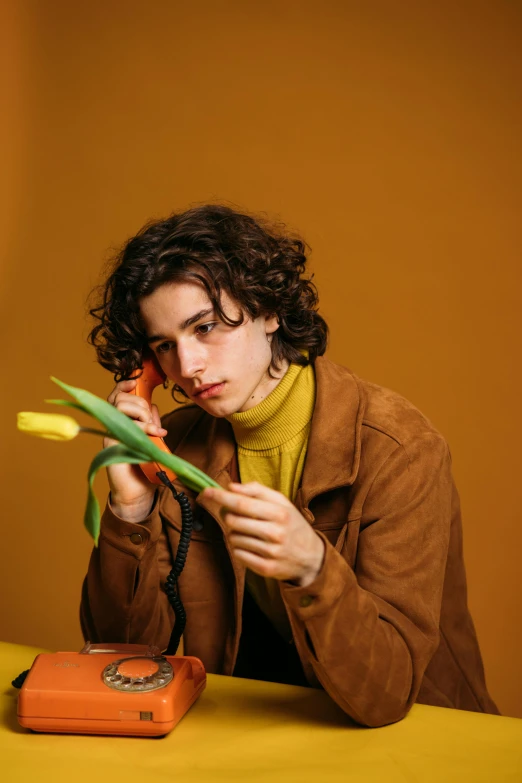 a man sitting at a table with a flower in his hand, an album cover, by Cosmo Alexander, trending on pexels, wavy hair yellow theme, attractive androgynous humanoid, timothee chalamet, greens)