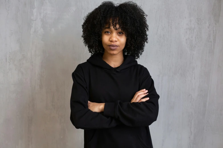 a woman in a black hoodie standing with her arms crossed, inspired by Afewerk Tekle, pexels contest winner, renaissance, curly bangs, professional headshot, panel of black, black girl