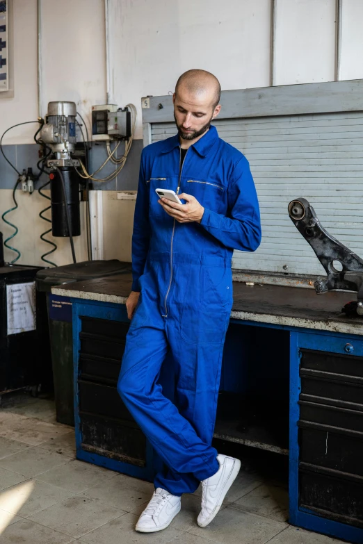 a man standing in a garage looking at his cell phone, inspired by Jan Müller, pexels contest winner, arbeitsrat für kunst, wearing human air force jumpsuit, kobalt blue, fullbody view, workbench