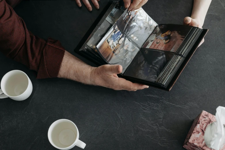 a couple of people that are holding a book, an album cover, unsplash, photorealism, on a coffee table, hasselblad photograph, contact sheet, high angle close up shot