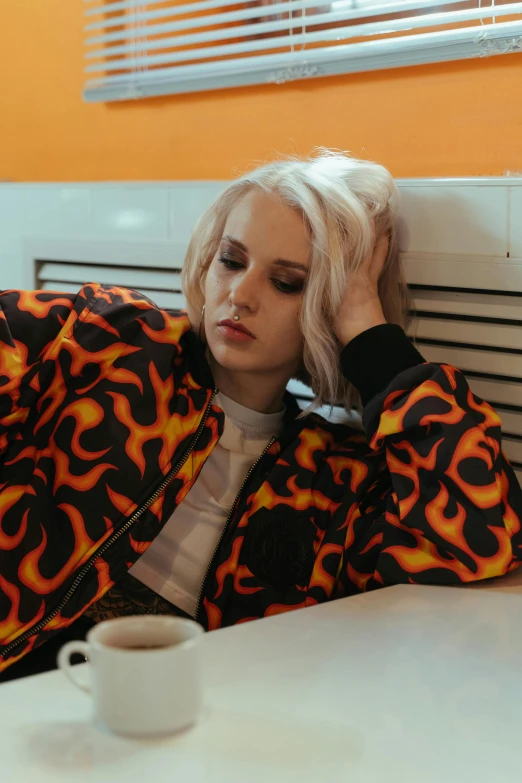 a woman sitting at a table with a cup of coffee, an album cover, inspired by Elsa Bleda, trending on pexels, graffiti, pale hair, black and orange coat, bedroom full of fire, pouting