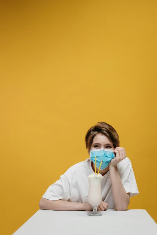 a woman sitting at a table with a face mask on, pexels contest winner, yellow backdrop, coronavirus, dentist, drinking