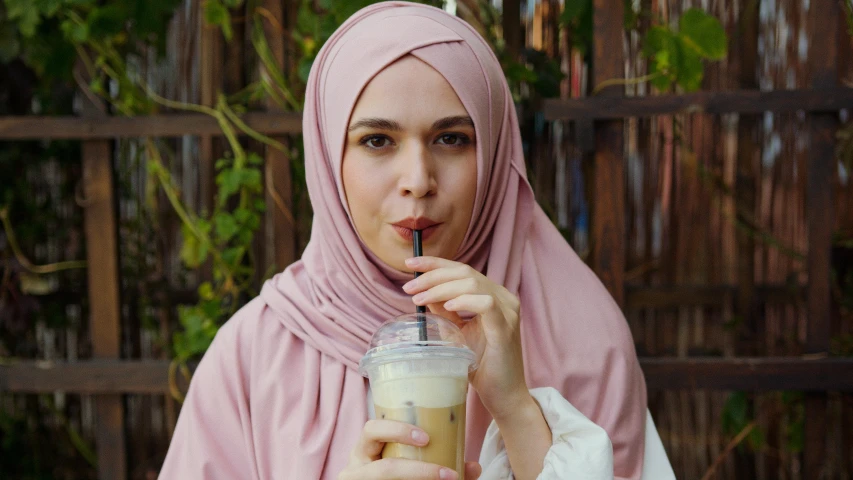 a woman in a hijab drinking a drink, inspired by Nazmi Ziya Güran, shutterstock, hurufiyya, iced latte, pink, her skin is light brown, pale thin lips