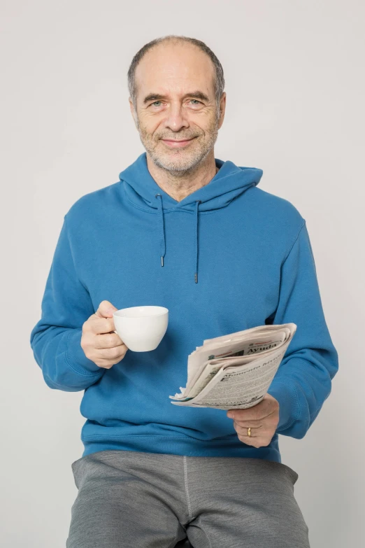 a man in a blue hoodie holding a newspaper and a cup of coffee, dark grey haired man, richard garriott, 2019 trending photo, studio portrait photo