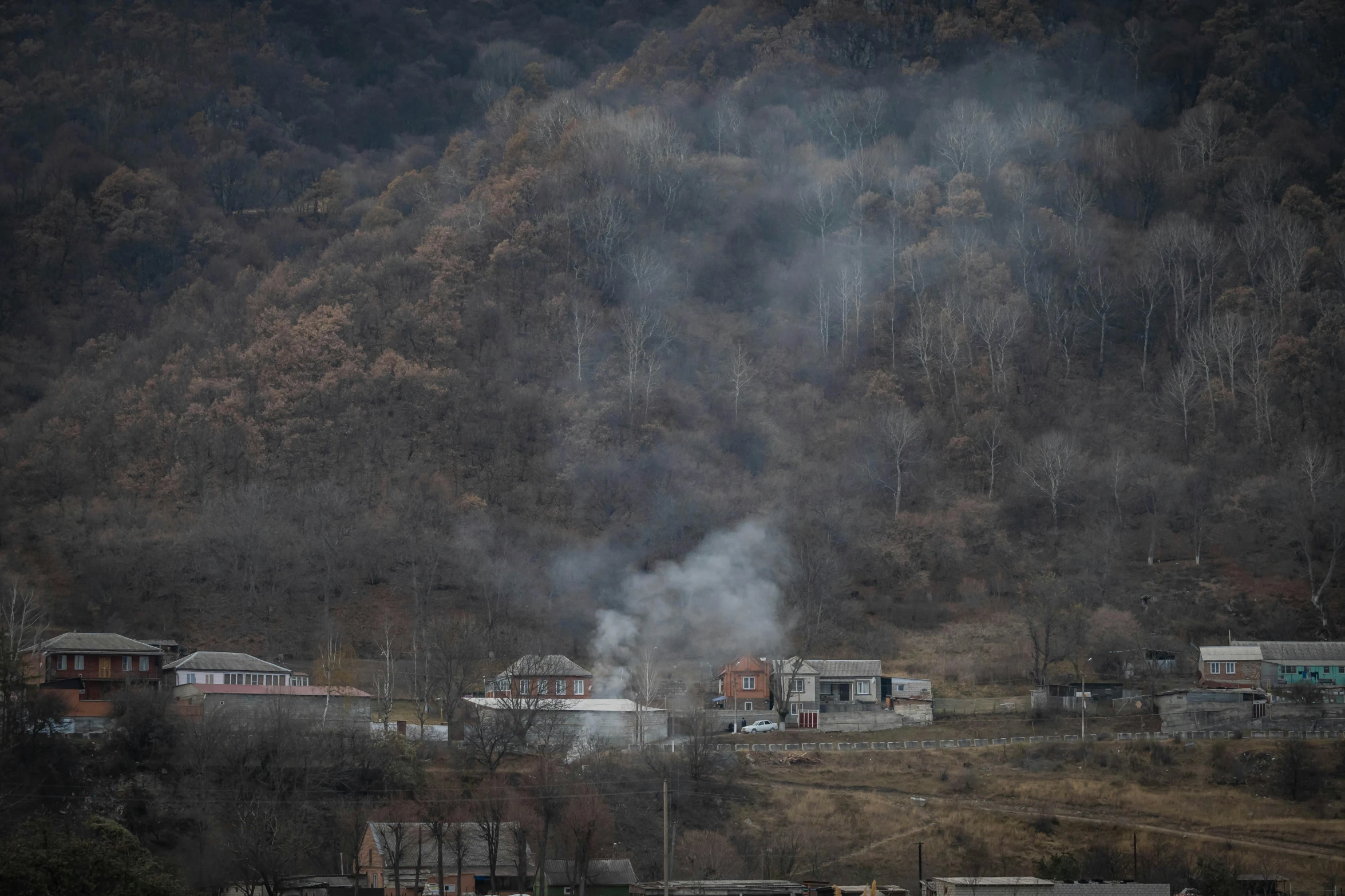 a small town with a lot of smoke coming out of it, by Muggur, hurufiyya, against the backdrop of trees, documentary photo, arma 3, shin jeongho