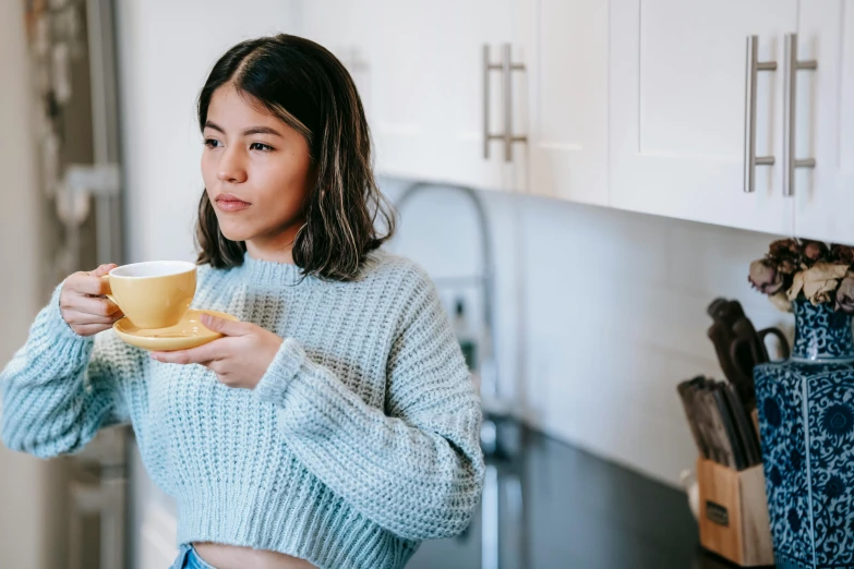 a woman holding a cup of coffee in a kitchen, trending on pexels, wearing yellow croptop, profile image, manuka, wearing casual sweater