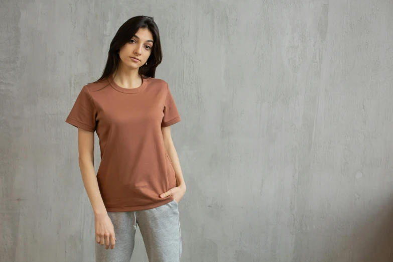 a woman standing in front of a concrete wall, trending on pexels, brown shirt, sport t-shirt, reddish - brown, small