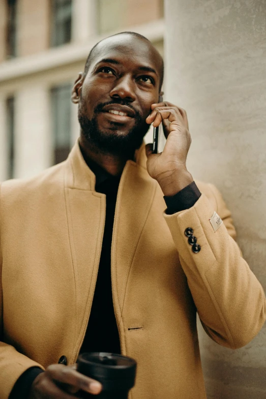 a man talking on a cell phone while holding a cup of coffee, by Daniel Gelon, trending on pexels, gold suit jacket in snake print, adut akech, high collar, muted brown yellow and blacks