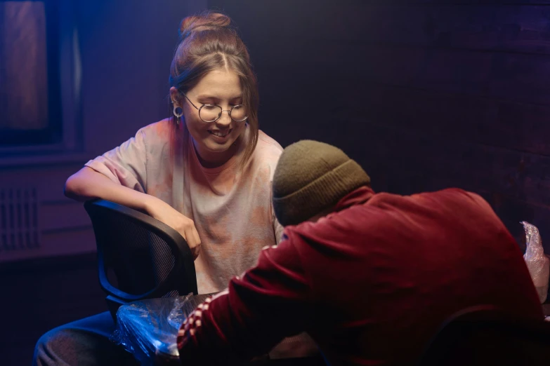 a woman sitting on top of a chair next to a man, rap scene, girl wearing round glasses, glowy light, worksafe. cinematic