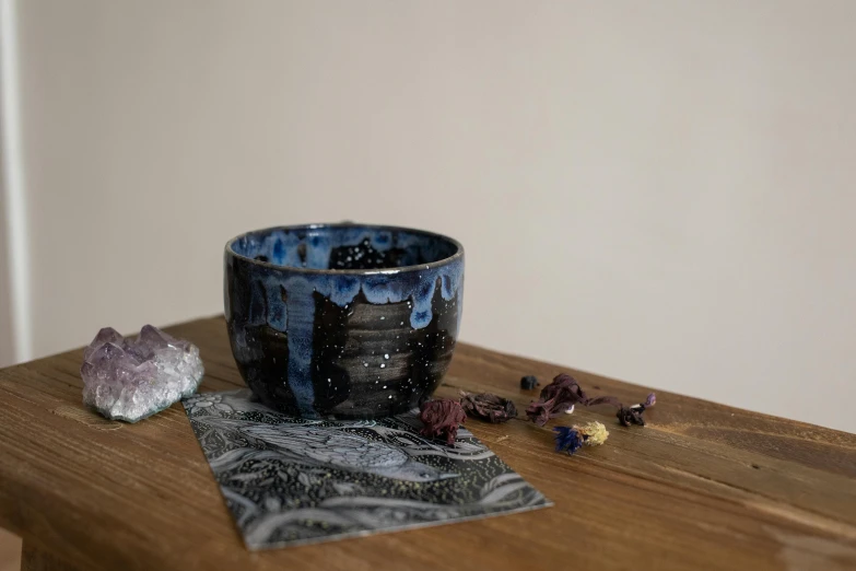 a black and blue bowl sitting on top of a wooden table, inspired by Hokusai, process art, crystals enlight the scene, black, labradorite, high detailed + tarot card