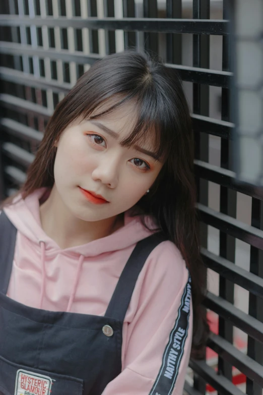 a woman in overalls leaning against a wall, a picture, inspired by Ma Yuanyu, trending on pexels, realism, wearing a pink hoodie, center parted bangs, vietnamese woman, square face