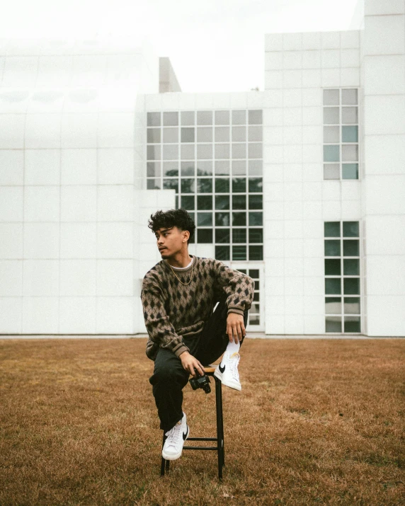 a man sitting on a chair in front of a building, an album cover, inspired by Russell Dongjun Lu, trending on unsplash, non binary model, grey sweater, posing for a picture, at the museum