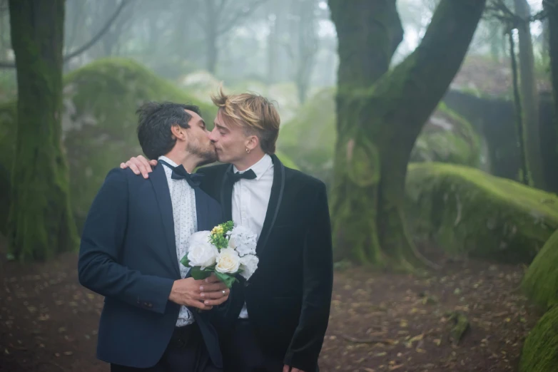 a couple of men standing next to each other in a forest, an album cover, by Romain brook, unsplash, romanticism, groom, kissing, youtube thumbnail, ignant