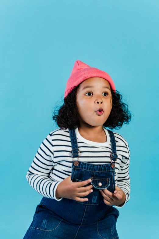 a little girl wearing a pink hat and overalls, pexels contest winner, pop art, surprised, blue backdrop, kodak color film, beanie