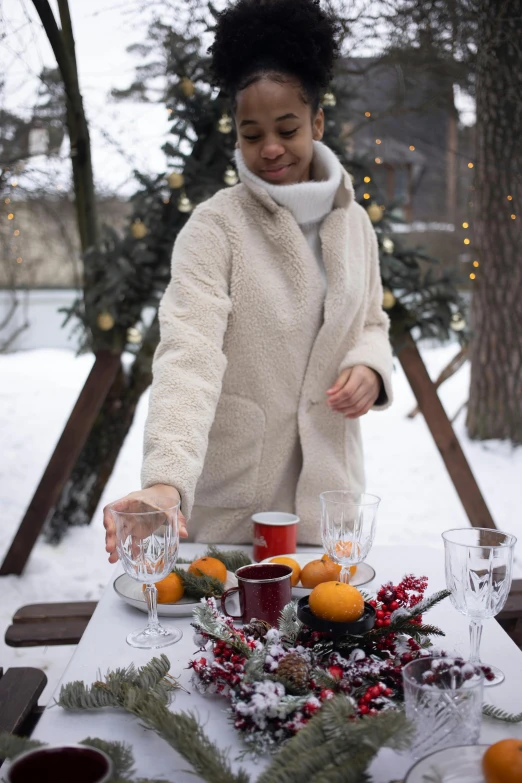 a woman standing in front of a table with oranges on it, winter wonderland, cheers, wearing hunter coat, white candles