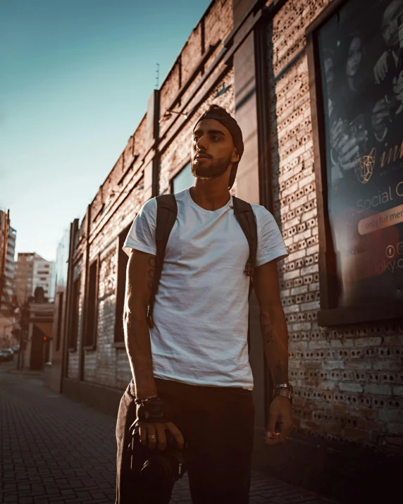 a man standing in front of a brick building, an album cover, pexels contest winner, man in white t - shirt, with a backpack, miguel iglesias, street pic