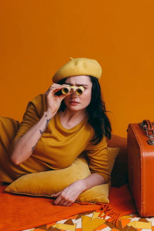 a woman sitting on a bed looking through a pair of binoculars, inspired by Wes Anderson, trending on pexels, orange and yellow costume, beret and sunglasses, in suitcase, charli xcx