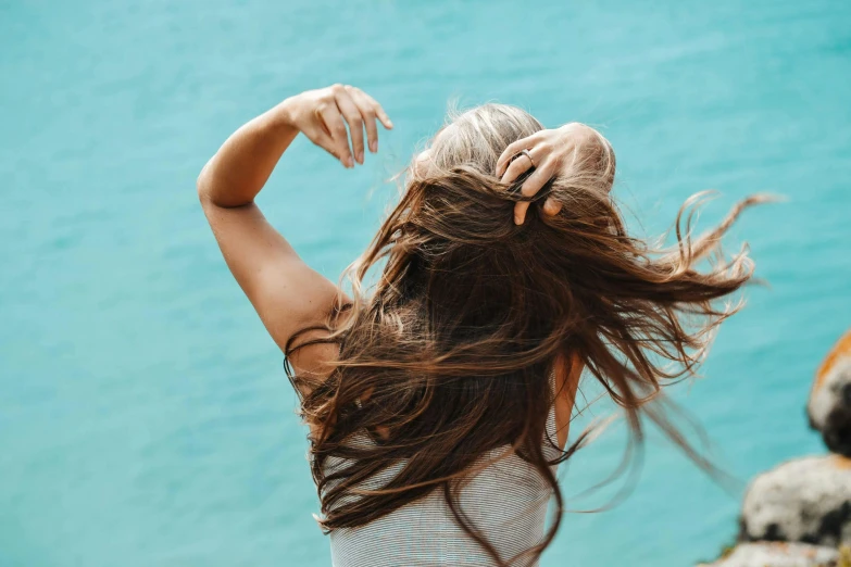 a woman standing on top of a cliff next to a body of water, trending on pexels, her hair is natural disheveled, she is dancing, with a blue background, hair : long brown