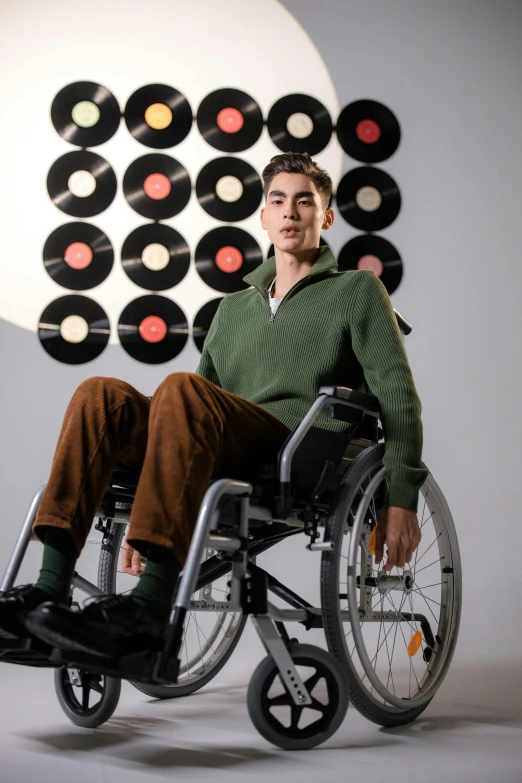 a man sitting in a wheel chair in front of a record wall, an album cover, inspired by Oskar Lüthy, in a dark green polo shirt, non binary model, he is wearing a brown sweater, ( ( theatrical ) )