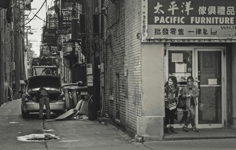 a couple of people standing in front of a building, a black and white photo, chinatown, poverty, parking in the street, “pig