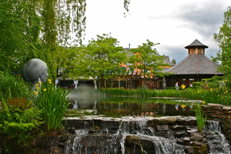 a small waterfall running through a lush green park, a picture, inspired by Matsumura Goshun, unsplash, hurufiyya, restaurant in background, moomin, viking palace, featuring flowing fountains