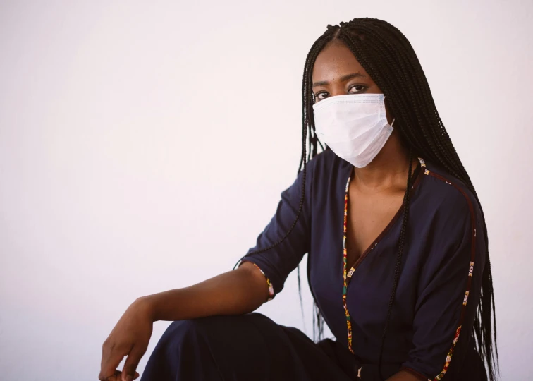 a woman wearing a face mask sitting on a chair, by Nina Hamnett, trending on pexels, afrofuturism, doctor, in front of white back drop, black female, dusty air