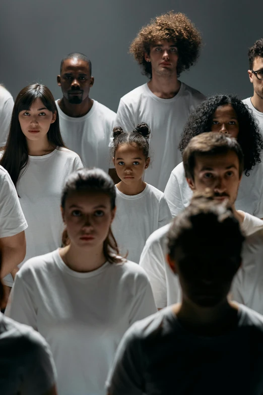 a group of people standing next to each other, dressed in a white t shirt, focused stare, dark. no text, diverse colors