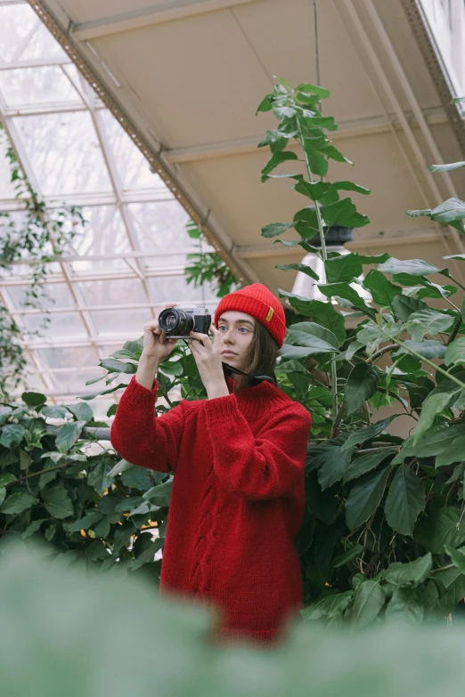 a woman taking a picture with a camera in a greenhouse, inspired by Elsa Bleda, visual art, wearing a red turtleneck sweater, red cap with a capital m, natalia dyer, botanical garden