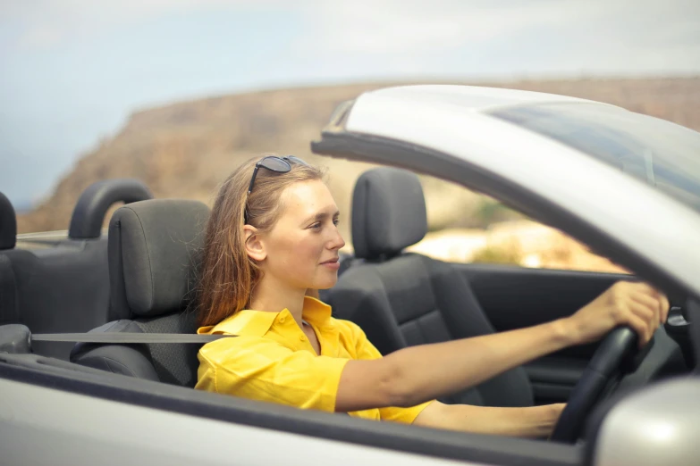 a woman sitting in the driver's seat of a convertible car, avatar image, square, yellow, left - hand drive