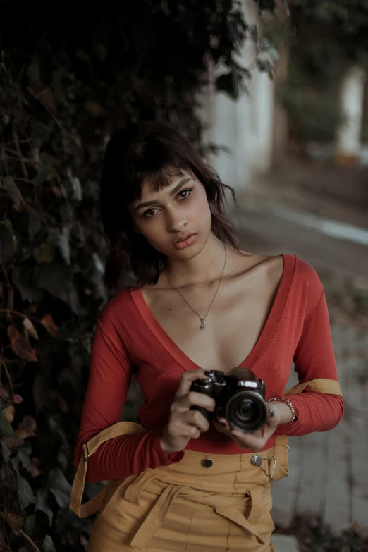 a woman standing next to a tree holding a camera, wearing red tank top, indian girl with brown skin, alessio albi, wearing a red outfit