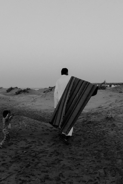 a man standing on top of a sandy beach, a black and white photo, by Ahmed Yacoubi, tattered cloth robes, at dusk, walk in a funeral procession, covered with blanket