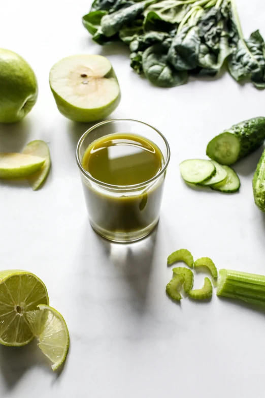 a table topped with green vegetables and a glass of juice, lime, goddess shot, oils, apple