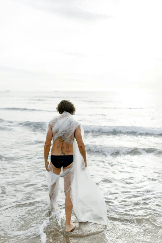 a man standing on top of a beach next to the ocean, an album cover, unsplash, renaissance, white loincloth, mullet, net of being, peter hurley