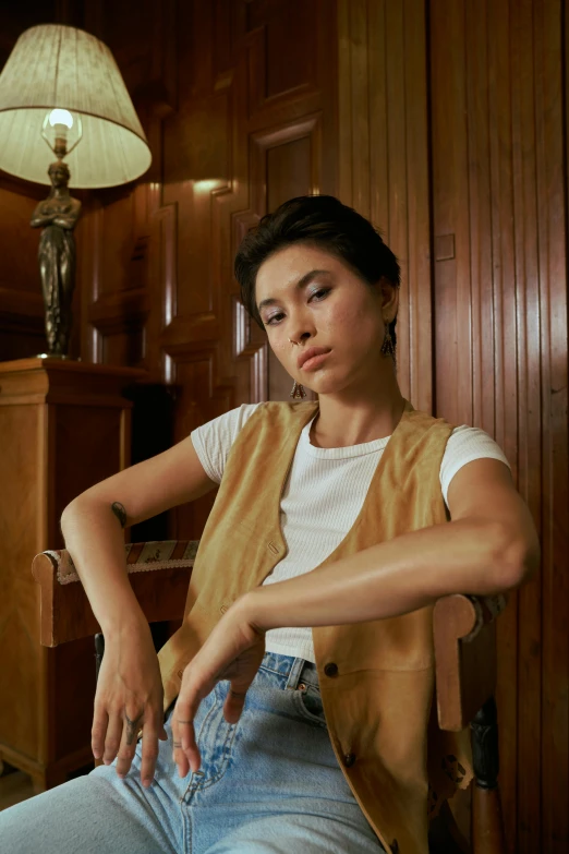 a woman sitting on a chair in a room, trending on pexels, photorealism, tan vest, portrait androgynous girl, cindy avelino, vintage aesthetic