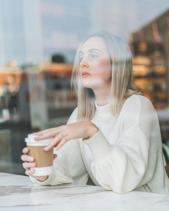 a woman sitting at a table with a cup of coffee, wearing a white sweater, looking through frosted glass, non-binary, wondering about others