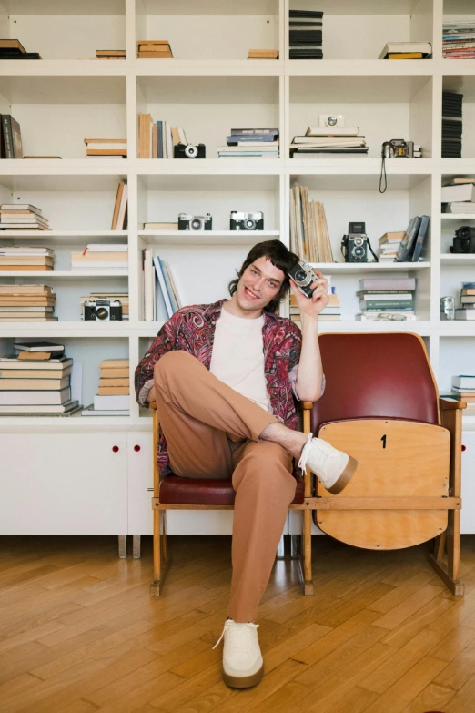 a man sitting in a chair in front of a bookshelf, an album cover, by Nina Hamnett, joe keery, high-quality photo, home album pocket camera photo, looking happy