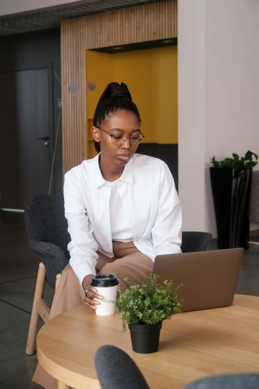 a woman sitting at a table with a laptop, wearing a white button up shirt, dark skinned, on a coffee table, next to a plant