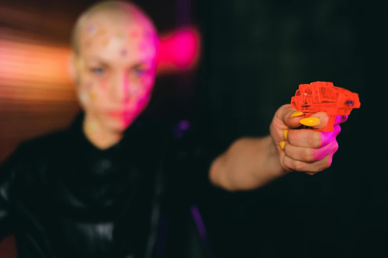 a close up of a person holding a toy gun, interactive art, shaved head, night clubs and neons, bella poarch, blurry