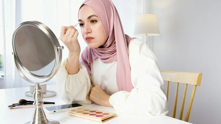 a woman sitting at a table in front of a mirror, inspired by Nazmi Ziya Güran, trending on pexels, hurufiyya, eyeshadow, white hijab, pink hue, easy to use